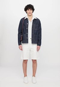 Denim skjortejakke med kontraststing, hvit skjorte under, hvite shorts og joggesko. Brun crossover-veske henger over skulderen.