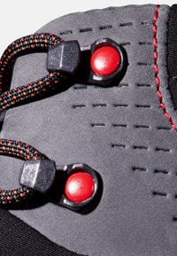 Czarne trampki z bliska, z teksturowanym materiałem, pomarańczowo-czarnymi sznurówkami, czerwonymi akcentami metalowymi oraz perforowanymi elementami wzorniczymi.