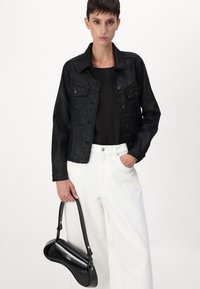 Giacca di jeans nera con tasche sul petto, abbinata a una maglia nera e pantaloni bianchi a gamba larga. Una borsa nera lucida è tenuta di lato.