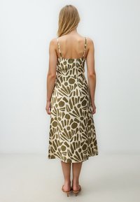 Maxi jurk van een beige stof met gedurfde groene abstracte patronen, dunne bandjes en een aansluitende top, die overgaat in een losse A-lijn rok.