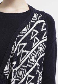 Stickad marinblå tröja med vitt geometriskt mönster framtill; har en v-ringning och mjuk textur, designad för en avslappnad passform.