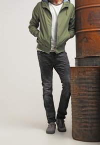 Veste verte à fermeture éclair avec capuche portée sur une chemise blanche, associée à un jean foncé et des baskets grises, debout à côté d'un baril rouillé.