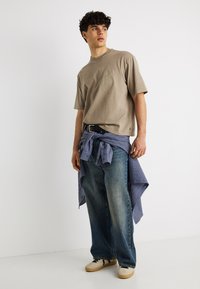 Νεαρός άνδρας που φοράει ένα μπεζ oversized t-shirt, μπλε τζιν, μπεζ αθλητικά παπούτσια και ένα μπλε πουκάμισο με καρό δεμένο γύρω από τη μέση του.