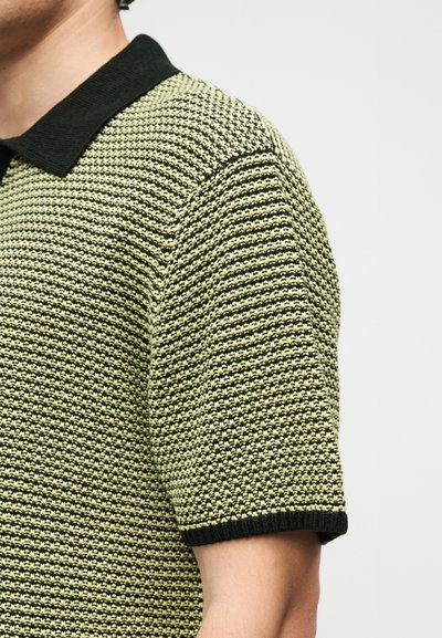 Κοντινό πλάνο ατόμου που φορά κοντομάνικο πλεκτό πόλο με μαύρο και κίτρινο-πράσινο υφασμάτινο μοτίβο και μονόχρωμο μαύρο γιακά και άκρα μανικιών.