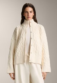 Kremasto barvan pulover s pletenim vzorcem, z visokim ovratnikom in zapenjanjem na gumbe. Rebrasti robovi na spodnjem delu in manšetah, z izrazitim vzorcem po celotni površini.