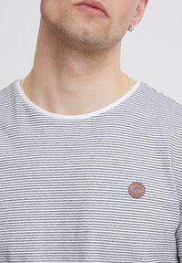 Prugasta majica s kratkim rukavima u bijeloj i tamnoplavoj boji, s okruglim izrezom i smešnim kožnim logom na prsima.