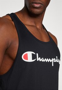 Canotta nera in cotone con scollatura rotonda. Presenta un logo "Champion" bianco con un accento rosso. Inquadratura ravvicinata della parte superiore del torso.