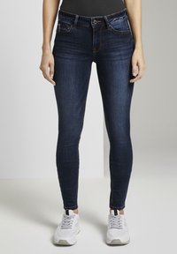 Jeans ajustados en un lavado azul oscuro, hechos de denim con costuras sutiles. Cuenta con cinco bolsillos y un cierre de cremallera, combinados con zapatillas blancas.