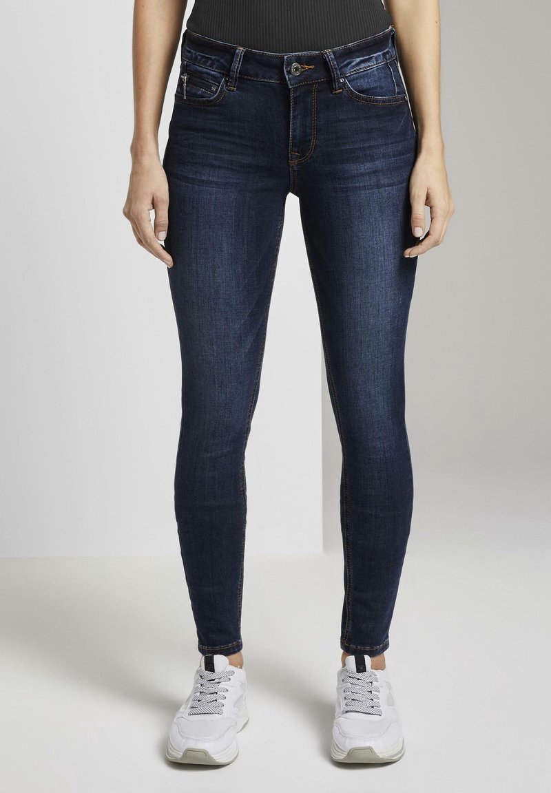 Jeans ajustados en un lavado azul oscuro, hechos de denim con costuras sutiles. Cuenta con cinco bolsillos y un cierre de cremallera, combinados con zapatillas blancas.