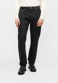 Μαύρο τζιν με ίσια γραμμή, διαθέτει μπροστινές τσέπες και κλείσιμο με κουμπιά. Φορεμένο με μαύρα παπούτσια χωρίς κορδόνια.