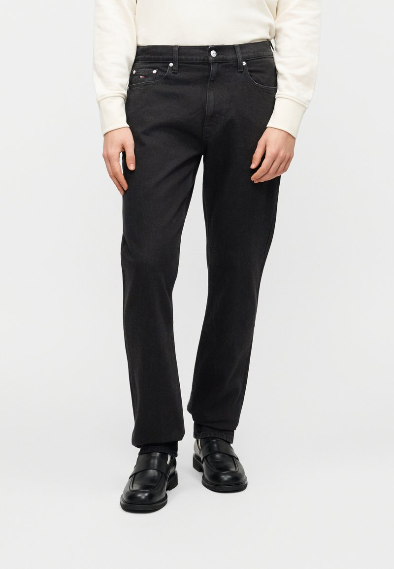 Μαύρο τζιν με ίσια γραμμή, διαθέτει μπροστινές τσέπες και κλείσιμο με κουμπιά. Φορεμένο με μαύρα παπούτσια χωρίς κορδόνια.