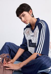Tumši zils un balts Adidas krekls ar mežģīņu paneli un logotipu, saskaņots ar tumši zilo svītraino biksēm. Modelis tur brūnu aukliņu somu.