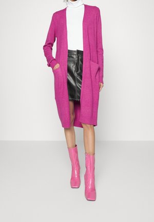 Frau trägt eine leuchtend pinke lange Strickjacke, schwarzen Lederrock, weißen Rollkragenpullover und glänzende pinke Stiefeletten, steht vor einem einfarbigen Hintergrund.