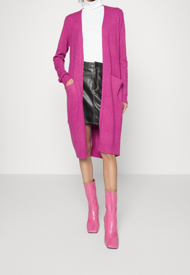 Femme portant un long cardigan rose vif, une jupe en cuir noire, un col roulé blanc et des bottines roses brillantes, debout devant un fond uni.