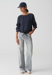 Maglietta a maniche lunghe blu navy con orlo regolabile, jeans larghi grigi chiari, sneakers bianche e berretto blu navy. Design minimalista e tessuti morbidi.