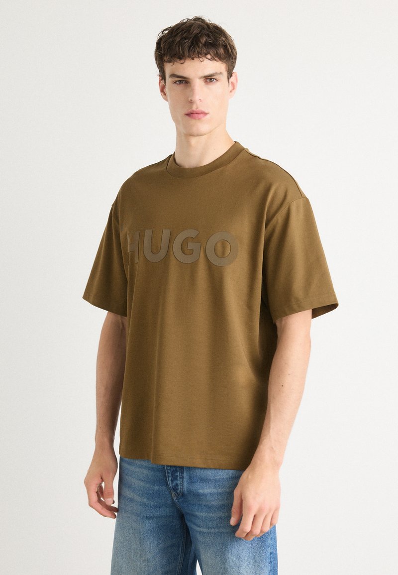 Pruun puuvillane T-särk ümmarguse kaelusega ja lühikeste varrukatega. Üksikasjana on prominentne "HUGO" logo veidi tumedamas tooni.
