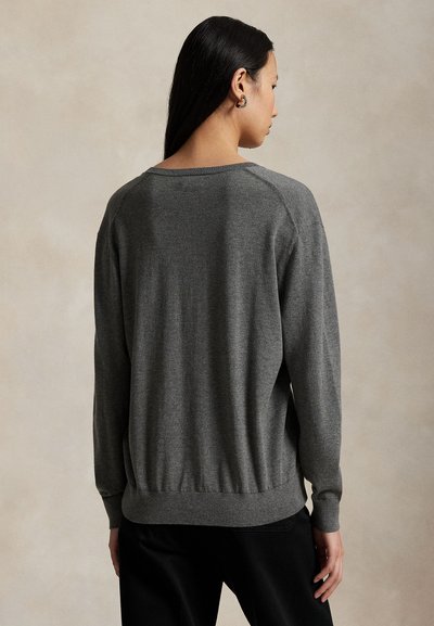 Moteris su ilgais tamsiais plaukais, dėvinti pilką megztinį ilgomis rankovėmis ir juodas kelnes, matoma iš nugaros neutraliame fone.