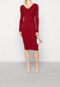 Robe rouge côtelée, jusqu'aux genoux, avec un décolleté en V et des manches longues, assortie à un petit sac à main beige et des talons beiges à lanières.