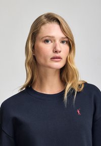 Marinblå sweatshirt med rundhals, mjuk känsla och en liten röd logotyp på vänster bröst. Designad för avslappnad användning.