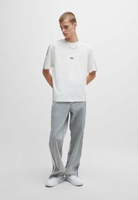 Weißes kurzärmliges T-Shirt mit blauem Logo, kombiniert mit hellgrauen Straight-Leg-Jeans mit blauen Seitenstreifen und weißen Sneakers.