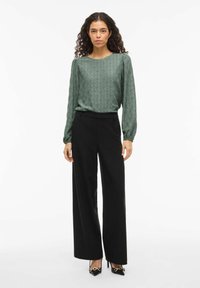 Zelená vzorovaná blůza s dlouhým rukávem, spárovaná s černými kalhotami s vysokým pasem a širokými nohavicemi, a černými lodičkami se zlatými detaily.