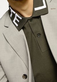 Šedý blazer s hladkou texturou a kulatým knoflíkem, spárovaný s tmavě olivovým polo tričkem s kontrastním bílým límečkem a detailním logem.