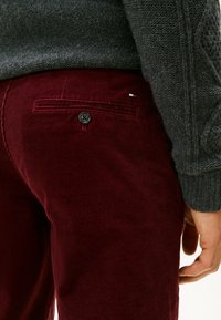 Bordeaux corduroy broek met een achterzaksluiting voorzien van een zwarte knop en een kleine logo-accent, gecombineerd met een grijze gebreide trui.