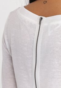 Maglia bianca in maglia con dettaglio della zip sul retro in metallo nero, scollo rotondo e vestibilità rilassata. La texture è leggera e trasparente.