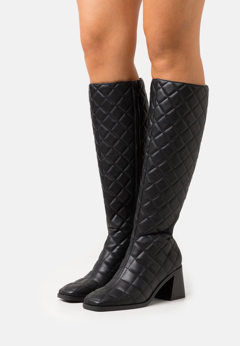 Bottes noires montantes en simili cuir matelassé, avec un bout carré et un talon épais, dotées d'une fermeture éclair sur le côté.