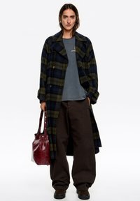 Długi płaszcz w kratę w ciemnej zieleni i granacie, gładka tkanina, zapięcie dwurzędowe. W połączeniu z szarą koszulą, ciemnymi, szerokimi spodniami oraz czerwonym torebką.