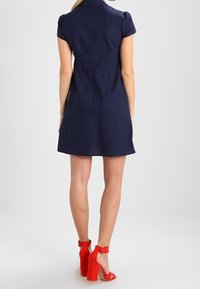 Tmavě modré šaty s krátkými rukávy, límečkem a přiléhavým pasem, mírně se rozšiřujícími do délky ke kolenům. Doplněné jasně červenými sandály na vysokém podpatku.