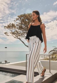 Haut noir sans manches avec un col froncé ; pantalon large rayé blanc avec un lien de serrage ; sandales ; arrière-plan extérieur pittoresque.