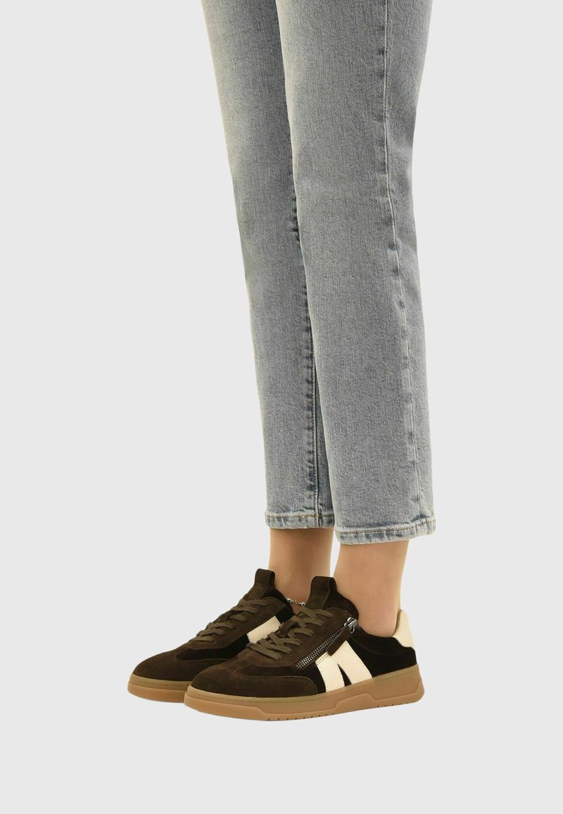Personne portant un jean bleu clair et des baskets marron avec des accents beiges et des fermetures éclair latérales, debout sur un fond neutre.