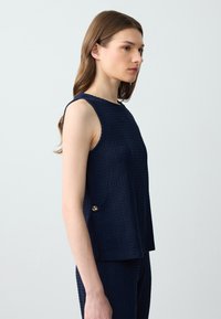 Femme aux longs cheveux bruns portant un haut sans manches texturé bleu marine et un pantalon assorti, se tenant de profil contre un fond clair.
