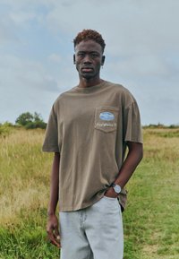 Jeune homme aux cheveux courts et bouclés, portant un t-shirt marron et un jean clair, debout dans un champ herbeux sous un ciel nuageux.