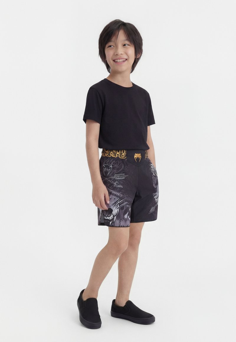 Ένα χαμογελαστό αγόρι στέκεται στραμμένο προς τα αριστερά, φορώντας μαύρο T-shirt, μαύρα γραφικά σορτς με κίτρινη μέση και μαύρα παπούτσια τύπου slip-on, πάνω σε λευκό φόντο.