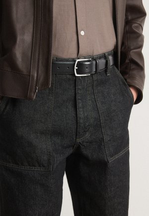 Black denim pants with contrasting yellow stitching, high waistband, and side pockets; paired with a brown leather jacket and striped shirt.