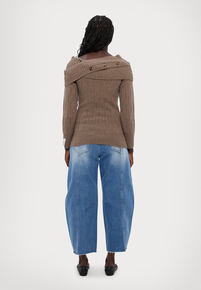 Brauner gerippter Strickpullover mit schulterfreiem Design und dekorativen Knöpfen, kombiniert mit lockeren blauen Jeans. Schwarze Schuhe runden den Look ab.
