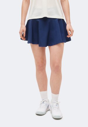 Person iført en mørkeblå plisseret nederdel, hvide sneakers, hvide crew-sokker og en hvid skjorte, stående mod en ensfarvet lys baggrund.