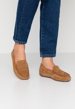 Mocassins en daim marron portés avec un jean en denim bleu, montrant un design à coupe basse et un détail de couture texturé sur un sol gris lisse.