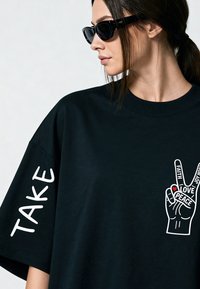 Femme portant des lunettes de soleil noires et un t-shirt noir avec un graphisme de main de paix blanche et le texte "TAKE" sur la manche, regardant sur le côté.