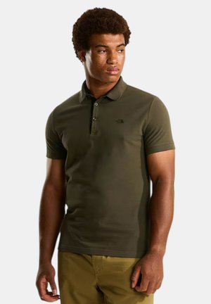 Joven vestido con una camisa tipo polo de manga corta color verde oliva y pantalones color caqui, de pie y mirando hacia su izquierda contra un fondo liso.