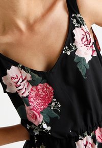 Černé květinové šaty s výstřihem do V, zdobené růžovými a červenými růžemi a zelenými listy. Materiál působí lehce a má jemnou texturu.