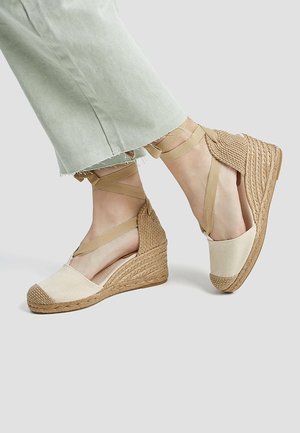 Beige lærred wedgesandaler med flettet jute-sål, ankelremme og en farveblokket tåcap. Lysegrønne, slidte bukser er delvist synlige.