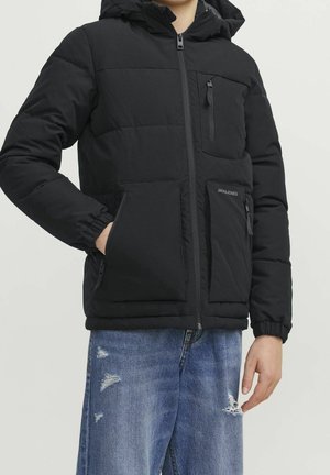 Personne portant une veste rembourrée noire avec des poignets élastiques, trois poches avant zippées, et un jean bleu déchiré, debout devant un fond uni.