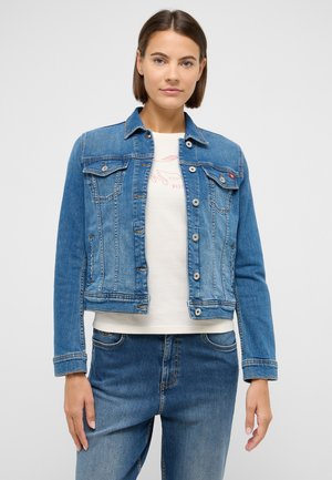 Young woman wearing a blue buttoned denim jacket over a white graphic T-shirt and blue jeans, standing against a plain light background.