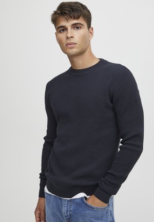 Pulover din tricot de tip waffle de culoare navy, cu guler rotund și mâneci lungi. Are manșete și tiv cu nervuri, purtat peste un tricou alb.