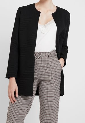 Femme portant une veste noire texturée ouverte, un caraco blanc orné de dentelle et un pantalon taille haute à carreaux avec une ceinture assortie.