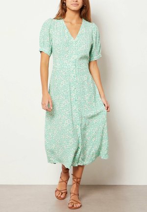 Femme portant une robe midi fleurie vert clair avec des manches courtes bouffantes et un décolleté en V, associée à des sandales plates marron à lacets.