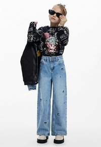 Top negru cu mâneci lungi cu design din paiete, blugi largi bleu deschis cu accente imprimate, jachetă neagră matlasată și ochelari de soare negri.